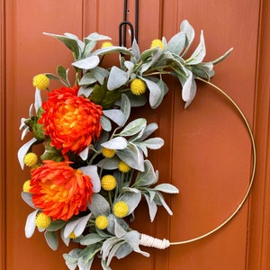 May include: A gold metal wreath with orange and yellow flowers and gray-green leaves. The wreath is hanging on a brown door.