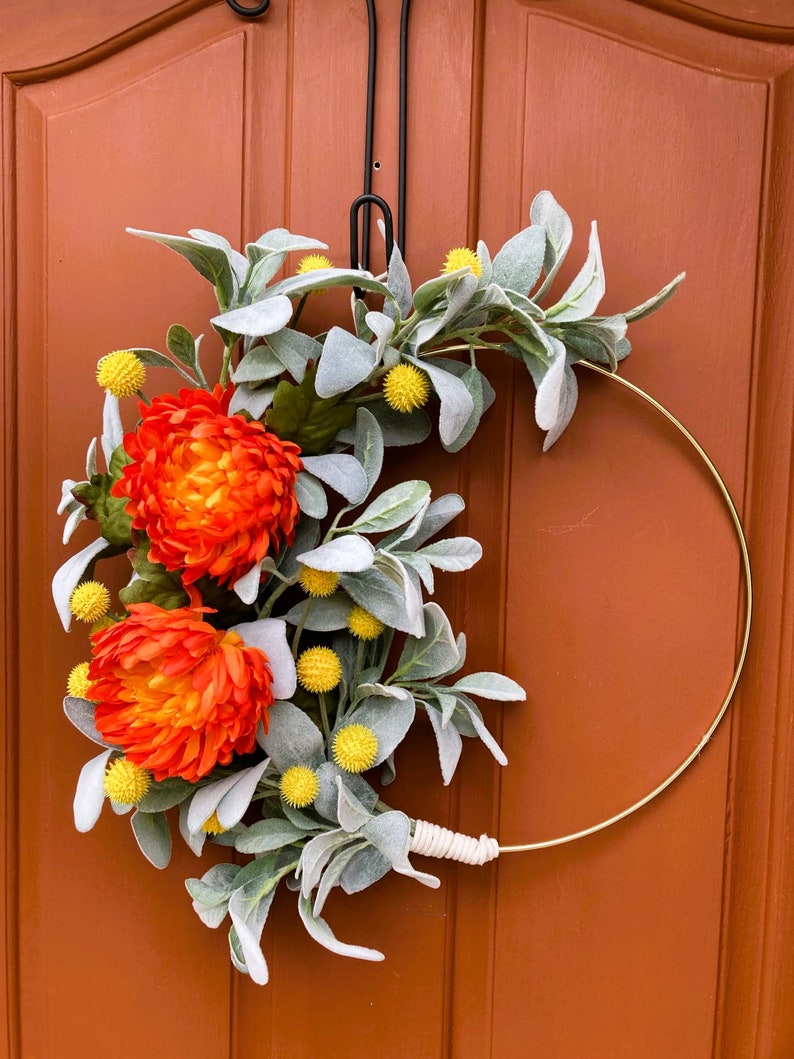 May include: A gold metal wreath with orange and yellow flowers and gray-green leaves. The wreath is hanging on a brown door.