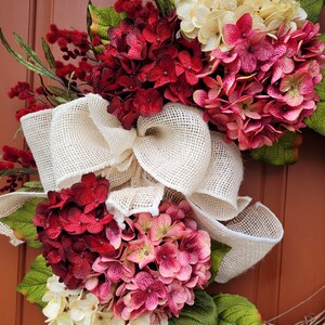Puede incluir: Una corona con un lazo de arpillera y tres hortensias de diferentes colores. Las hortensias son blancas, rosadas y rojas.