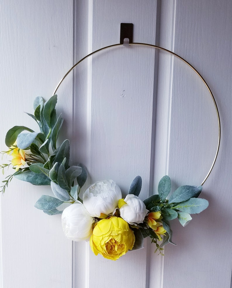May include: A gold metal wreath with white and yellow flowers and green leaves. The wreath is hanging on a white door.