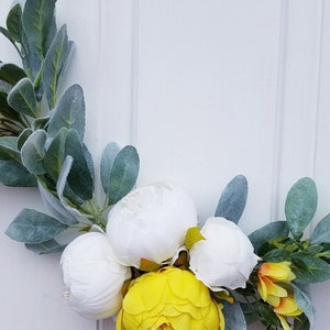 May include: A wreath made with white and yellow flowers and green leaves. The wreath is on a white background.