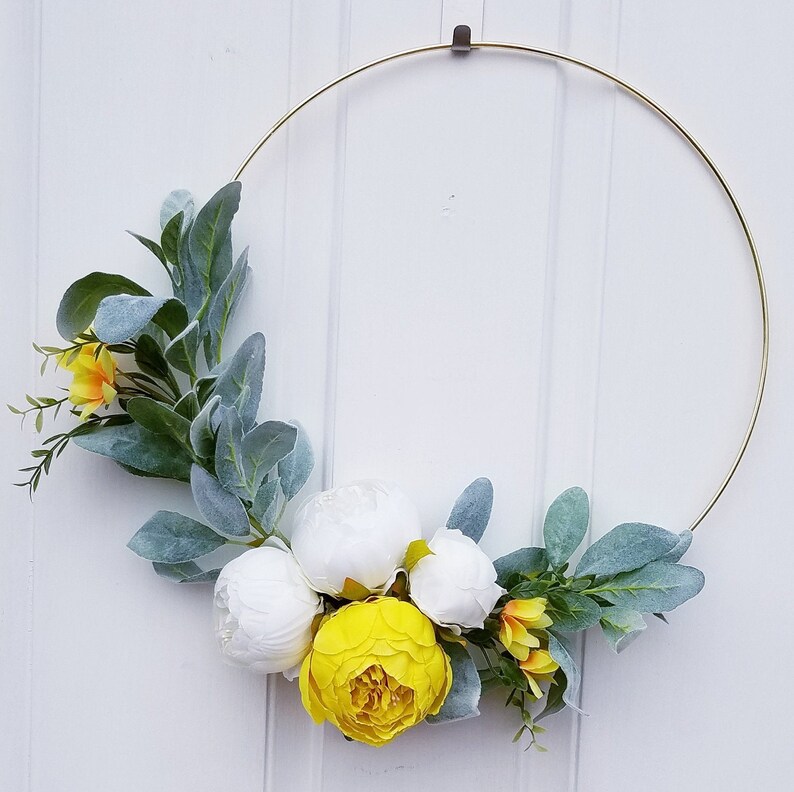 May include: A gold metal wreath with white and yellow flowers and green leaves. The wreath is hanging on a white wall.