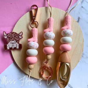 Pode incluir: Três chaveiros de contas rosa e brancas com um amuleto de bota de cowboy rosa. O chaveiro à esquerda tem um aro de chave dourado. O chaveiro do meio tem um aro de chave dourado e uma conta de madeira. O chaveiro à direita tem uma conta de madeira e um difusor de garrafa dourado. Os chaveiros estão sobre uma superfície de madeira com um fundo rosa. O texto "Miss Flora & Co." é visível no canto inferior esquerdo da imagem.