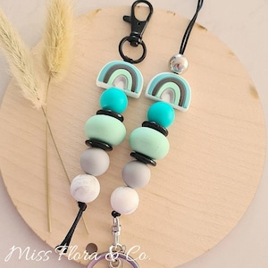 Pode incluir: Um chaveiro com um amuleto de arco-íris, um anel de chave prateado e um cordão preto. O amuleto apresenta um design de arco-íris com cores verde, branco e cinza. O chaveiro também tem uma série de contas coloridas em tons de cinza, verde menta e turquesa.
