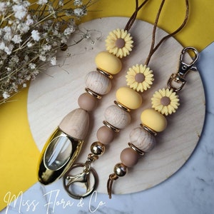 Pode incluir: Um chaveiro de contas amarelas e marrons com um anel de chave de metal dourado e uma pequena garrafa de vidro com uma tampa dourada. As contas são feitas de madeira e silicone e apresentam desenhos de girassóis.