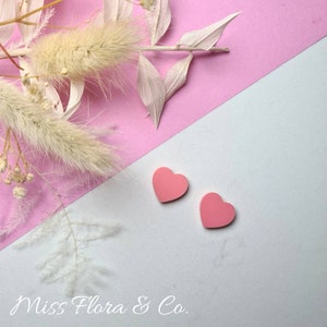 Pu&ograve; includere: Due orecchini a forma di cuore rosa su uno sfondo bianco e rosa. Lo sfondo &egrave; diviso in diagonale, la met&agrave; superiore &egrave; rosa e la met&agrave; inferiore bianca. Gli orecchini sono nell'angolo in basso a destra dell'immagine. Il testo "Miss Flora & Co." &egrave; nell'angolo in basso a sinistra dell'immagine.