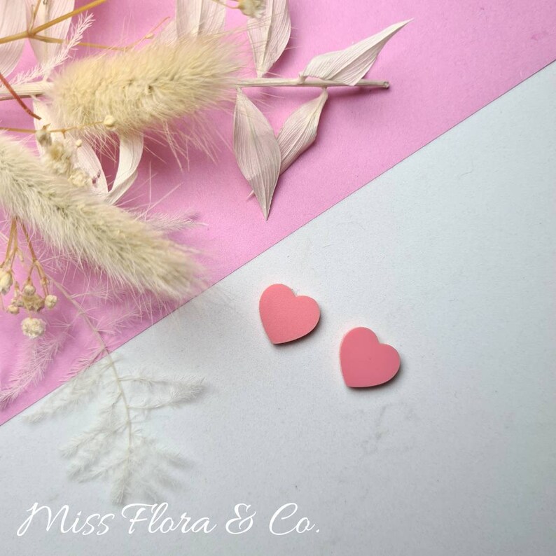 Pu&ograve; includere: Due orecchini a forma di cuore rosa su uno sfondo bianco e rosa. Lo sfondo &egrave; diviso in diagonale, la met&agrave; superiore &egrave; rosa e la met&agrave; inferiore bianca. Gli orecchini sono nell'angolo in basso a destra dell'immagine. Il testo "Miss Flora & Co." &egrave; nell'angolo in basso a sinistra dell'immagine.