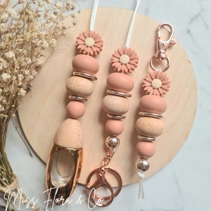 May include: Three beaded lanyards with a daisy accent, wooden beads, and rose gold hardware. One lanyard has a rose gold key fob and a wooden perfume bottle. The lanyards are displayed on a wooden circle.