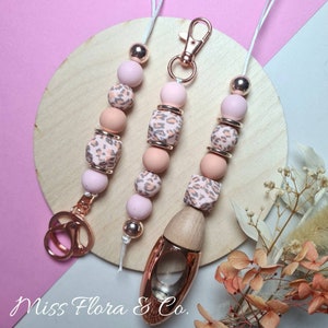 Pode incluir: Três chaveiros de contas cor-de-rosa e castanhas com detalhes dourados. O chaveiro à direita tem uma garrafa difusora de madeira anexada. Os chaveiros estão sobre uma superfície de madeira castanho claro. "Miss Flora & Co." está escrito em texto preto sobre a superfície.