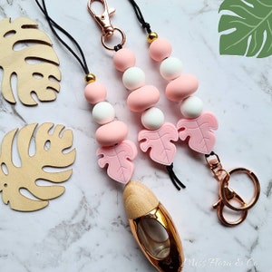 Könnte beinhalten: Drei Perlen-Lanyards mit rosa, weißen und goldenen Akzenten. Jedes Lanyard hat einen rosa Blatt-Charm und einen roségoldenen Verschluss. Ein goldener und hölzerner Diffusor für ätherische Öle ist ebenfalls sichtbar. Die Lanyards befinden sich auf einer Marmoroberfläche.