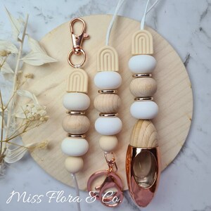 Pode incluir: Três aromatizadores de carro com um design de contas de madeira e um amuleto de silicone. Os amuletos têm a forma de um arco. Os aromatizadores estão presos a um chaveiro de ouro rosa. Os aromatizadores estão sobre uma superfície de madeira. "Miss Flora & Co."