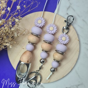 May include: Three lavender and wood beaded keychains with a flower charm. Each keychain has a silver ring and a silver clip. The keychains are on a wooden surface.