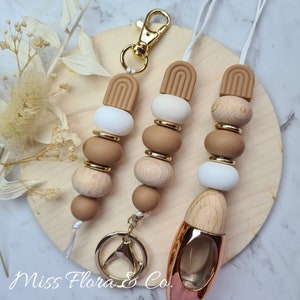 Pode incluir: Três aromatizadores de carro com um chaveiro dourado. Cada aromatizador tem uma corrente de contas marrons e brancas com um acento dourado. Os aromatizadores têm a forma de um arco marrom e branco. Os aromatizadores estão sobre uma superfície de madeira.