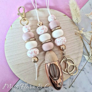 May include: Three pink and white beaded keychains with gold accents and a gold key ring. The keychains are hanging from a wooden surface. A small, clear glass bottle with a gold top and a wooden cap is also on the surface. The bottle is a diffuser.