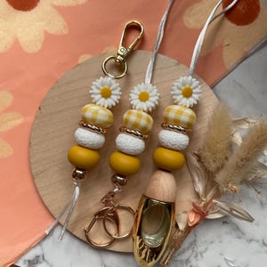 Könnte beinhalten: Drei Perlen-Lanyards mit Gänseblümchen- und Vichy-Akzenten. Die Lanyards haben gelbe, weiße und goldfarbene Perlen mit einem goldfarbenen Verschluss und Schlüsselring. Der Hintergrund umfasst einen Holzring und florale Elemente.