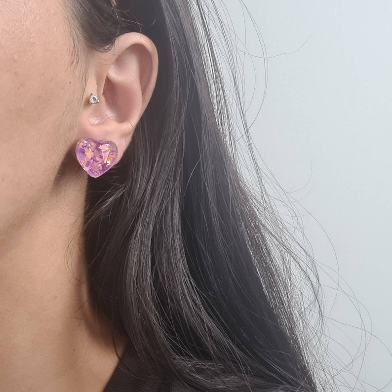 Pu&ograve; includere: Un paio di orecchini a forma di cuore con una finitura iridescente viola e rosa. Gli orecchini sono indossati nell'orecchio sinistro della persona.