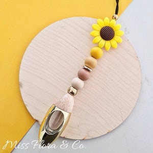 Pode incluir: Um ambientador decorativo para carro com um design de girassol. O ambientador apresenta um girassol amarelo, contas de madeira de várias cores e um difusor de metal e madeira dourados. O texto "Miss Flora & Co." é visível.