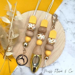 Könnte beinhalten: Ein Set aus drei gelben und weißen Perlen-Auto-Lufterfrischern mit einem goldenen Schlüsselanhänger und einer goldenen Diffuserflasche. Die Perlen haben ein Sonnenblumenmuster. Die Diffuserflasche ist mit einer klaren Flüssigkeit gefüllt. Der Text "Miss Flora & Co." ist in der rechten unteren Ecke des Bildes sichtbar.