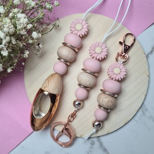 Pode incluir: Uma coleção de cordões e chaveiros de contas rosa e madeira. Os cordões apresentam detalhes de margaridas rosa, contas de madeira e anéis de metal em ouro rosa. Um chaveiro tem um pingente em forma de carro em ouro rosa.