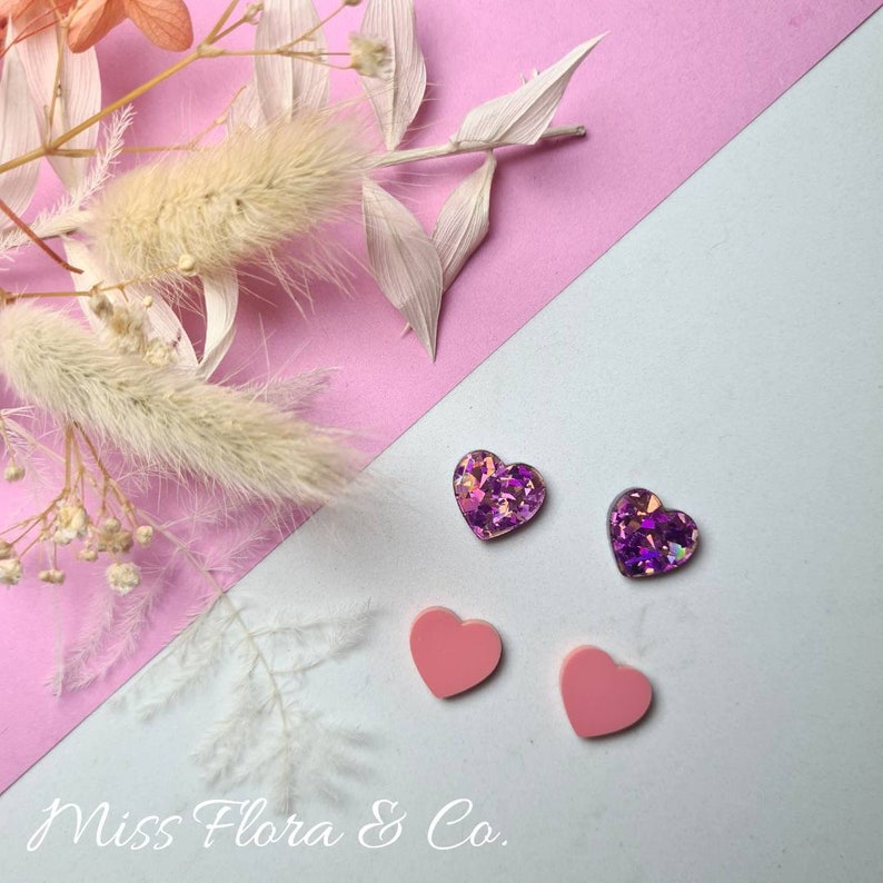 Pu&ograve; includere: Quattro orecchini a forma di cuore, due rosa e due viola con glitter. Gli orecchini sono su uno sfondo rosa e bianco. Miss Flora & Co.