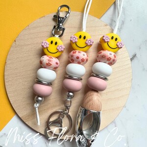 Pode incluir: Três chaveiros com contas rosa e brancas e um amuleto de rosto sorridente amarelo com flores rosa. Os chaveiros estão presos a um chaveiro prateado, um difusor prateado e de madeira e um cordão branco.