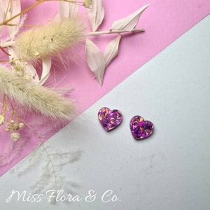 Pu&ograve; includere: Un paio di orecchini a forma di cuore con una finitura glitter rosa e viola. Gli orecchini sono piccoli e delicati, perfetti per aggiungere un tocco di brillantezza a qualsiasi outfit.