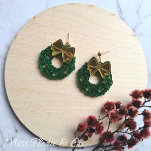 May include: A pair of green glitter Christmas wreath earrings with gold bows. The earrings are on a wooden surface.