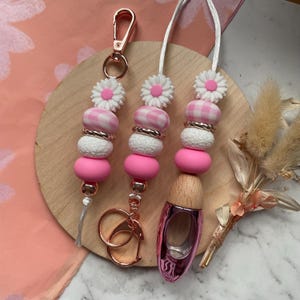Puede incluir: Tres llaveros con cuentas y un cordón con un tema de margarita rosa y blanco. Los llaveros presentan cuentas rosas, blancas y de cuadros vichy con detalles dorados. Un llavero tiene un cierre y anillos de oro rosa. El cordón tiene un dije en forma de zapato rosa.