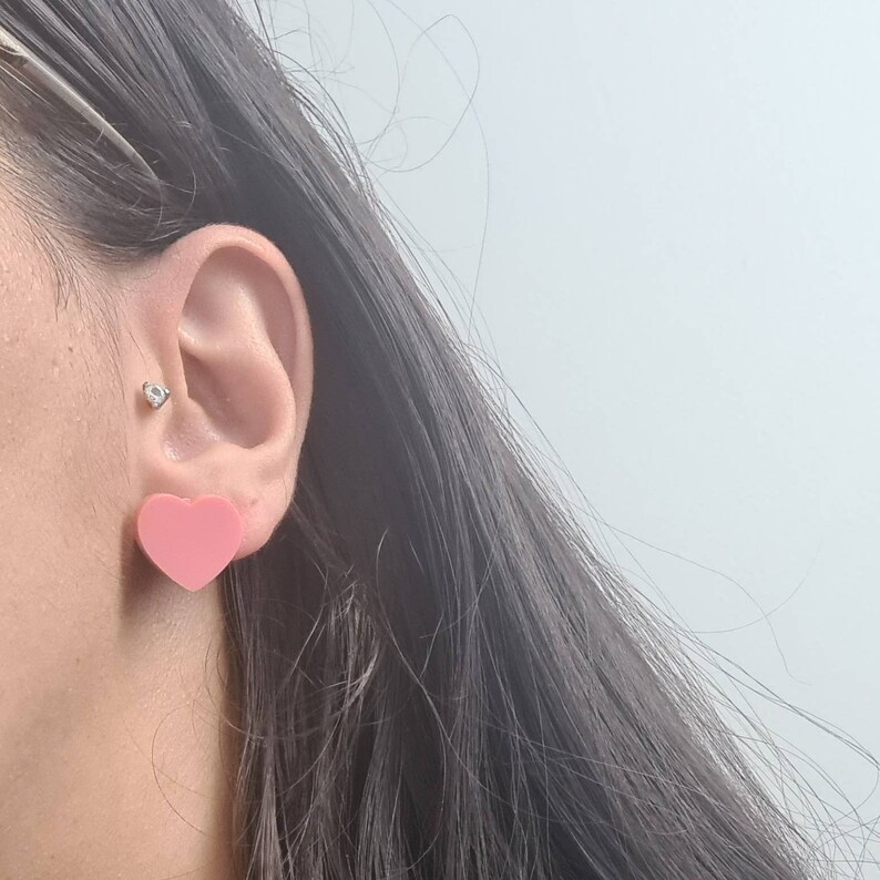 Pu&ograve; includere: Primo piano di un orecchio con un orecchino a forma di cuore rosa. L'orecchino &egrave; realizzato in un materiale liscio e lucido.