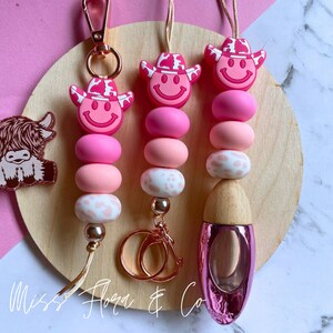 Pode incluir: Três chaveiros de contas rosa e brancas com chapéus de caubói e carinhas sorridentes. Os chaveiros estão presos a argolas douradas e a uma garrafa de vidro rosa com tampa de madeira. O texto "Miss Flora & Co" é visível no canto inferior esquerdo da imagem.