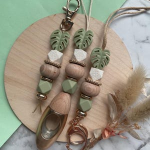 Pode incluir: Chaveiros com mosquetão dourado e ambientador de carro dourado. Os chaveiros apresentam contas verdes, brancas e de madeira, além de pingentes em forma de folha verde. Sobre uma superfície de madeira.