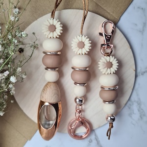 Pode incluir: Três acessórios de contas com detalhes de margaridas. Os acessórios apresentam uma combinação de contas creme, castanhas e prateadas. Um tem uma garrafa cor de ouro rosa, e outro tem um fecho cor de ouro rosa. Os acessórios são enfiados em cordões castanhos.