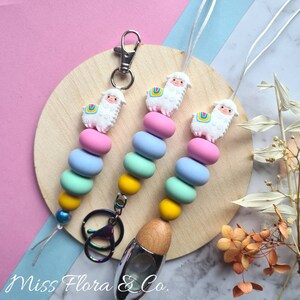 Pode incluir: Três chaveiros de contas coloridas com um amuleto de lhama. Cada chaveiro tem um esquema de cores diferente: rosa, azul e verde. Os chaveiros estão sobre uma superfície de madeira com um fundo branco. O texto "Miss Flora & Co." é visível no canto inferior esquerdo da imagem.