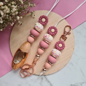 May include: A set of three pink and white beaded keychains with floral charms. Each keychain features a different pattern of pink and white beads, including a floral pattern and a speckled pattern. The keychains are attached to a gold-coloured metal ring and a gold-coloured metal clip.