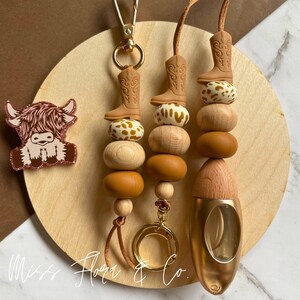 Pode incluir: Coleção de chaveiros e acessórios artesanais. Os chaveiros apresentam uma bota de cowboy, contas de madeira e um fecho dourado. Um alfinete de vaca Highland rosa e castanho também está incluído. O texto "Miss Flora & Co." é visível.