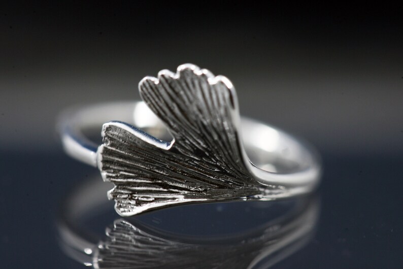 K&ouml;nnte beinhalten: Ein silberner Ring mit einem detaillierten Ginkgoblatt-Design. Das Blatt ist gebogen und scheint aus Metall zu sein.