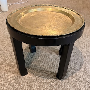 May include: A round side table featuring a black wooden base with four curved legs and a removable circular brass tray top. The brass tray has a scalloped edge and intricate engraved patterns across its surface, providing a metallic contrast.