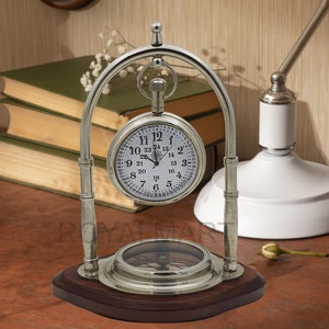 May include: A silver pocket watch with a compass on a wooden stand. The watch face has black Roman numeral hour markers and black minute markers. The compass has a green needle and a white face with black markings.