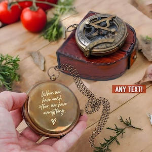 Może przedstawiać: Okrągły mosiężny kompas kieszonkowy z łańcuszkiem, zawierający wygrawerowany tekst "When we have each other, we have everything." oraz symbol serca. Drugi mosiężny kompas słoneczny spoczywa na ciemnobrązowym sk&oacute;rzanym etui.