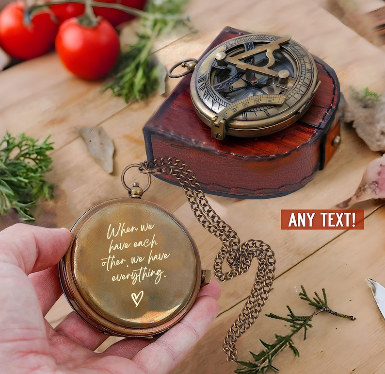 Może przedstawiać: Okrągły mosiężny kompas kieszonkowy z łańcuszkiem, zawierający wygrawerowany tekst "When we have each other, we have everything." oraz symbol serca. Drugi mosiężny kompas słoneczny spoczywa na ciemnobrązowym sk&oacute;rzanym etui.