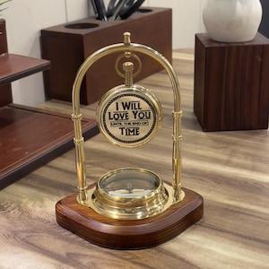 May include: A brass desk ornament featuring a circular compass base mounted on a dark wood stand. A brass arch supports a circular plaque with the engraved text I WILL LOVE YOU UNTIL THE END OF TIME, suspended above the compass by a small ring.