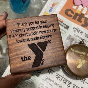 Może przedstawiać: Kwadratowe drewniane pudełko o ciemnobrązowym wykończeniu z wygrawerowanym laserowo tekstem: Thank you for your visionary support in helping the Y chart a bold new course towards north Eugene, wraz z logo YMCA oraz słowem the.