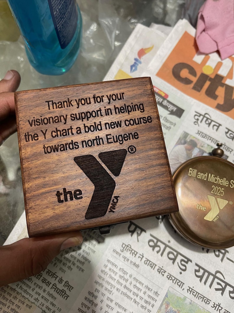 Może przedstawiać: Kwadratowe drewniane pudełko o ciemnobrązowym wykończeniu z wygrawerowanym laserowo tekstem: Thank you for your visionary support in helping the Y chart a bold new course towards north Eugene, wraz z logo YMCA oraz słowem the.