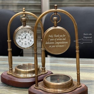 May include: A brass clock and compass set on a wooden base. The clock face has Roman numerals and the compass has a red needle. The clock is engraved with the text "Andy, Thank you for 17 years of service and dedication. Congratulations on your retirement!!" and the back side is engraved.