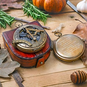 May include: A vintage brass compass with a leather case sits next to a round brass compass with engraved text. The compasses are on a wooden surface with fall foliage and pumpkins in the background. The text on the round compass reads: "A compass guides the adventurer."