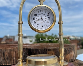 May include: A brass pocket watch with Roman numeral markings on a white face is displayed on a brass stand with a wooden base. The watch face also has a compass rose design. The stand has a decorative finial on top. The text "Royalraonadeem.etsy.com" is visible on the base.