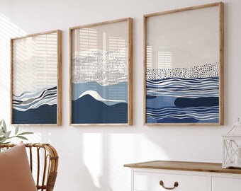 Puede incluir: Tres impresiones de arte abstracto enmarcadas con olas azules y blancas sobre un fondo crema. Las impresiones están colgadas en una pared blanca sobre una cómoda blanca con una tapa de madera. Una silla de mimbre con un cojín marrón y una manta blanca está en primer plano.