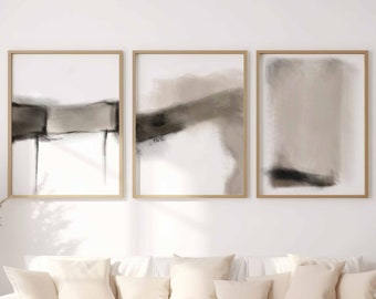 May include: A white sofa with beige pillows sits in front of a white wall with three framed abstract paintings in shades of brown and gray. A wooden coffee table with a tray, two mugs, and a stack of books sits in front of the sofa.