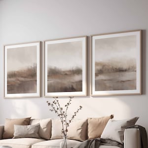 May include: A light beige sofa with three cushions and a throw blanket, two wooden coffee tables, and three framed abstract landscape paintings in muted brown tones on the wall behind the sofa.