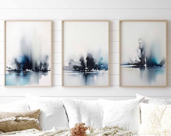 May include: Three abstract watercolour paintings in a white frame, hanging on a white wall above a white sofa with a throw blanket and cushions.